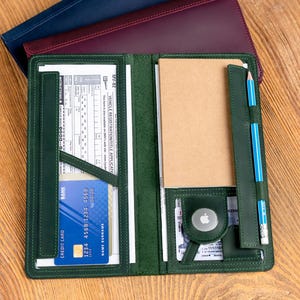 May include: A green leather folio with a credit card, notepad, and pencil. The folio is open, revealing interior pockets and a notepad. Two additional folios in navy and burgundy are stacked behind it on a wooden surface.