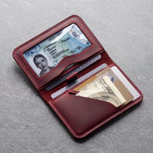 May include: Open burgundy leather wallet with a clear ID window displaying a driver's license. The wallet also holds credit cards and a folded US dollar bill. The wallet is open, revealing its interior and contents.