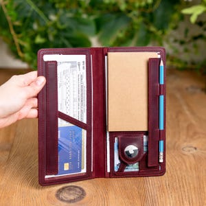May include: Open burgundy leather notebook with multiple pockets, holding a notepad, pencil, credit card, and documents. The notebook is open, revealing its interior. The background is a wooden surface.