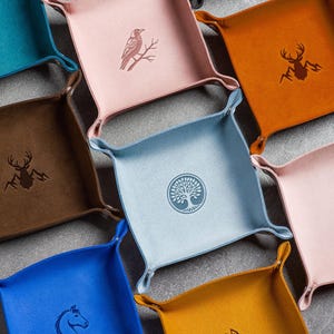 May include: A collection of square leather trays in various colors, including pink, blue, and orange. Each tray features a unique embossed design, such as a raven, deer, horse, wolf, and tree. The trays have curved edges and are arranged on a gray surface.