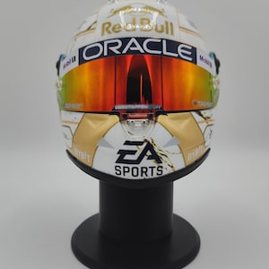 Puede incluir: Casco de carreras blanco y dorado con los logotipos de Red Bull y Oracle, y una visera espejada. El casco está sobre un soporte negro, con el logotipo de EA Sports. Artículo de colección.