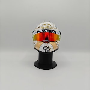 Peut inclure: Un casque de course blanc et doré arborant les logos Red Bull et Oracle, une visière orange réfléchissante et la marque EA Sports sur la mentonnière. Le casque est présenté sur un support noir, mettant en valeur les logos et graphismes.