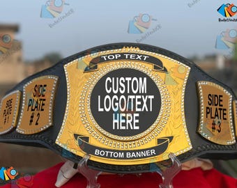 Puede incluir: Un cinturón de campeonato negro y dorado con el texto "TOP TEXT", "CUSTOM LOGO/TEXT HERE" y "BOTTOM BANNER". El cinturón tiene placas laterales con el texto "SIDE PLATE #1", "SIDE PLATE #2" y "SIDE PLATE #3".