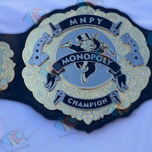 Könnte beinhalten: Ein gold-schwarzer Monopoly Champion-Gürtel. In der Mitte ist der Monopoly-Mann mit dem Wort "MONOPOLY" darüber und "CHAMPION" darunter abgebildet. Der Gürtel hat goldene Akzente und Würfel auf den Seitenteilen.