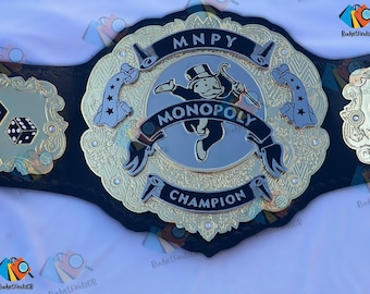 May include: A gold and black Monopoly Champion belt. The center features the Monopoly man with the word "MONOPOLY" above and "CHAMPION" below. The belt has gold accents and dice on the side plates.