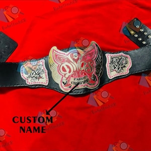 Könnte beinhalten: Ein schwarzer Leder-Wrestling-Meisterschaftsgürtel mit einem großen zentralen Emblem mit einem rosa-goldenen Design. Der Gürtel hat Seitenteile mit dem WWE-Logo und dem Text "Paris Champion".