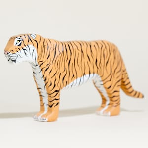 May include: A tiger figurine featuring a vibrant orange coat with distinct black stripes. The animal is depicted in a standing position, showcasing white fur details on the face, chest, and underbelly, with a long tail extending behind the body.