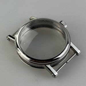 Könnte beinhalten: Ein silberfarbenes Uhrengehäuse mit einem transparenten Kristallzifferblatt. Das Gehäuse hat eine runde Form mit einer strukturierten Lünette und Bandanstößen zum Anbringen eines Armbands. Die Krone ist an der Seite sichtbar.