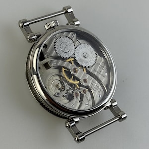 Puede incluir: Una vista de cerca de un reloj de pulsera plateado vintage, que revela su intrincado movimiento mecánico. La esfera del reloj muestra engranajes, resortes y el texto "South Bend Watch Co. USA". El reloj tiene barras plateadas para la correa.