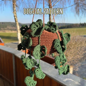 Könnte beinhalten: Ein hängender Häkeltopf mit einem braunen Topf und grünen Blattakzenten. Der Topf wird von braunen Kordeln gehalten und trägt den Text "DIGITAL PATTERN". Die Häkelarbeit ist detailliert und erzeugt ein strukturiertes Aussehen.