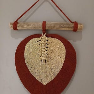 Peut inclure: Une décoration murale composée d'une grande pièce en macramé en forme de feuille rouge foncé, superposée à une pièce plus petite en macramé doré. L'ensemble est suspendu à un morceau de bois flotté clair par une cordelette rouge foncé.