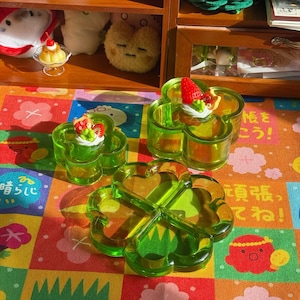 Könnte beinhalten: Drei kleeblattförmige, transparente grüne Glasschalen. Zwei Schalen haben ein kleines Dessert mit einer Erdbeere oben drauf. Die Schalen stehen auf einer bunten Oberfläche mit Blumenmuster. Im Hintergrund ein Holzregal mit Plüschtieren.