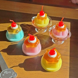 Op de afbeelding: Een verzameling kleurrijke, miniatuur jelly desserts met een kers erop. De desserts zijn in tinten geel, roze en blauw en staan op een houten oppervlak. Sommige staan op kleine glazen voetstukken. Een decoratieve lepel is zichtbaar.