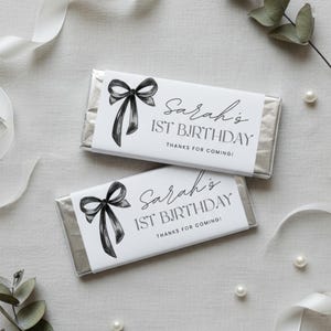 Op de afbeelding: Twee chocoladerepen verpakt in zilverfolie, elk met een wit etiket met een zwarte strik illustratie en de tekst "Sarah's 1st Birthday, Thanks for Coming!". Lint en parels op een lichte achtergrond.