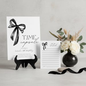 Puede incluir: Una tarjeta de cápsula del tiempo en blanco y negro con una ilustración de lazo, exhibida en un soporte negro. Una tarjeta más pequeña con un lazo y las palabras "Dear Sarah" está al lado. Un jarrón negro contiene rosas blancas y vegetación.