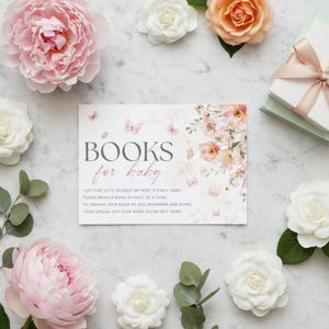 Puede incluir: Tarjeta de invitación a un baby shower con el texto "BOOKS for baby", rodeada de flores rosas y blancas y mariposas. La tarjeta tiene un diseño floral y una petición a los invitados para que traigan un libro en lugar de una tarjeta.