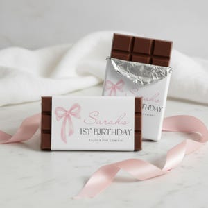 Op de afbeelding: Twee chocoladerepen met gepersonaliseerde verpakkingen. Eén reep is gedeeltelijk uitgepakt en toont de chocolade. De verpakkingen hebben een roze strik illustratie, de naam "Sarahs" en de tekst "1ST BIRTHDAY THANKS FOR COMING!" op een witte achtergrond.