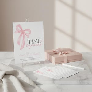 Puede incluir: Un conjunto de cápsula del tiempo rosa para un primer cumpleaños. El conjunto incluye una tarjeta con una ilustración de lazo rosa y las palabras "Time Capsule", una caja de regalo con una cinta rosa y dos tarjetas de notas. También se incluye un bolígrafo blanco.