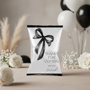 Könnte beinhalten: Weiße Party-Geschenktüte mit einer schwarzen Schleifenillustration und dem Text "THANKS FOR COMING WITH LOVE Sarah". Die Tüte ist von weißen und schwarzen Luftballons, weißen Rosen und dekorativen Pflanzen umgeben.