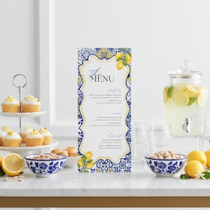 Könnte beinhalten: Eine Tischdekoration mit Speisekarte, Cupcakes und Limonade. Die Speisekarte hat ein blaues und gelbes Design mit Zitronen. Cupcakes stehen auf einem mehrstöckigen Ständer. Ein Glasgetränkespender enthält Limonade. Schalen mit blau-weißen Mustern sind mit Nüssen gefüllt.