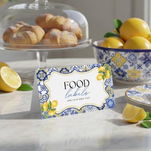 Può includere: Un'etichetta alimentare bianca con un design floreale blu e giallo e le parole "FOOD labels" in corsivo nero. L'etichetta ha un'illustrazione di limone e foglie. Una ciotola di limoni e croissant è sullo sfondo.