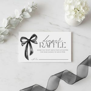 Może przedstawiać: Biały kupon na loterię baby shower z czarną ilustracją kokardy. Tekst brzmi "diaper RAFFLE" i "BRING THIS TICKET AND A PACK OF DIAPERS FOR YOUR CHANCE TO WIN A PRIZE". Czarna wstążka jest ułożona na dole.