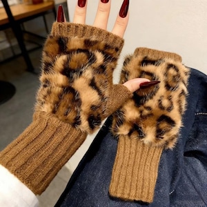 以下が含まれることがあります： 茶色のヒョウ柄のデザインの指なし手袋。手袋は茶色のニットカフスと、上部にフェイクファーのヒョウ柄プリントが施されています。手袋は人の手の上に表示されています。