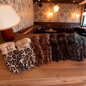以下が含まれることがあります： ヒョウ柄の手袋のコレクション。フェイクファーのトリムとバックルアクセントが特徴です。手袋は木製の表面に展示されており、さまざまな色とパターンが示されています。手袋は暖かさとスタイルを考慮して設計されています。
