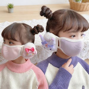 May include: Two children wearing decorative face masks. One mask is light pink with a plush character and a red bow. The other mask is lavender with a plush crown and purple bows. Both masks have white trim and ear coverings.