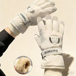 May include: White winter gloves with a dog face design on the back of the hand. The gloves have a wrist strap with the word "Pochacco" printed on it. The fingertips are gray and the inside is lined with a soft, brown material.