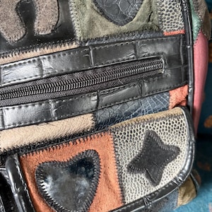 Puede incluir: Un bolso de mano de patchwork con diversas texturas y colores, incluyendo negro, marrón, verde y rosa. El bolso presenta formas de corazón y estrella cosidas, un bolsillo horizontal con cremallera y paneles con patrones de cocodrilo y granulados.