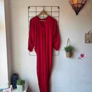 May include: A striking red dress with a V-neck and long, draped sleeves hangs on a wooden hanger. The dress is displayed against a white wall with a black grid and decorative elements, including a hanging lamp and small plants.