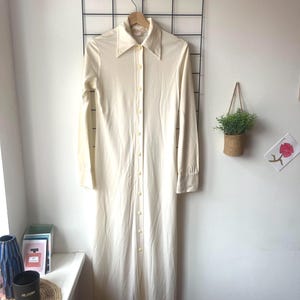 May include: A cream-coloured long dress with a collared neckline and long sleeves. The dress has a button-down front and hangs on a wooden hanger against a black grid. The dress is a vintage style.