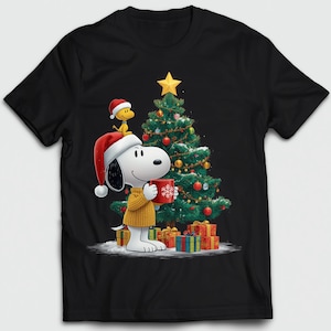 May include: Black t-shirt featuring Snoopy wearing a Santa hat and holding a red mug with a snowflake design. A Christmas tree with ornaments and a star is behind Snoopy. Woodstock is on Snoopy's head. Presents are at the base of the tree.