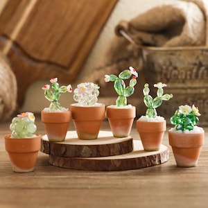 Op de afbeelding: Een collectie van zes decoratieve miniatuurcactussen gemaakt van groene en doorzichtige gefacetteerde glaskralen, elk geplant in een kleine terracotta pot gevuld met wit grind. De cactussen zijn voorzien van kleine witte en roze glazen bloemen.