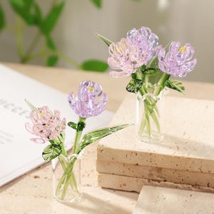 May include: Two glass flower sculptures, one pink and one purple, each in a small glass vase. The flowers have detailed petals and green stems and leaves. The vases sit on a light-colored, textured surface.