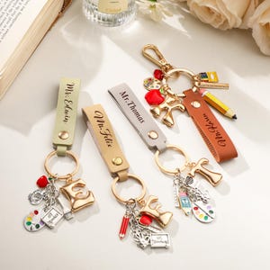 May include: Four personalised leather keychains in light green, tan, grey, and brown, each featuring a gold-tone metal ring, a letter charm, and miniature school-themed charms like a pencil, palette, and apple, with names engraved on the leather.