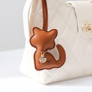 Könnte beinhalten: Ein brauner Leder-Katzen-Anhänger mit einer goldenen Kette und einem kleinen runden Anhänger mit der Aufschrift "Lucky girl". Der Anhänger hängt an einer weißen, gesteppten Handtasche mit goldenem Verschluss.