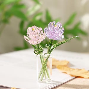 May include: Two glass flower sculptures, one pink and one purple, are displayed in a small glass vase. The flowers have detailed petals and green stems and leaves. The vase sits on a white surface with a blurred green background.