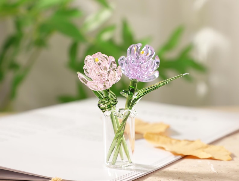 May include: Two glass flower sculptures, one pink and one purple, are displayed in a small glass vase. The flowers have detailed petals and green stems and leaves. The vase sits on a white surface with a blurred green background.