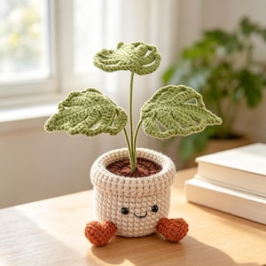 Puede incluir: Una planta de ganchillo con hojas verdes y una maceta sonriente. La planta tiene tres hojas verdes y está en una maceta de color crema con una cara sonriente. Dos pequeños corazones naranjas están colocados delante de la maceta.