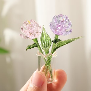 May include: A miniature glass flower arrangement featuring two detailed blooms in pink and purple, with green stems and leaves. The flowers are held in a small clear glass vase, creating a delicate and artistic display.