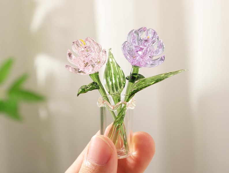 May include: A miniature glass flower arrangement featuring two detailed blooms in pink and purple, with green stems and leaves. The flowers are held in a small clear glass vase, creating a delicate and artistic display.