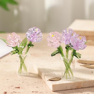 May include: Two miniature glass flower arrangements. One features pink and purple flowers, the other has pink and purple flowers. Each arrangement is in a small clear glass vase with green stems and leaves. The flowers have yellow centers.
