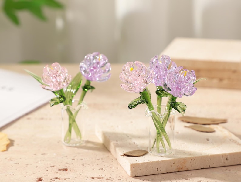May include: Two miniature glass flower arrangements. One features pink and purple flowers, the other has pink and purple flowers. Each arrangement is in a small clear glass vase with green stems and leaves. The flowers have yellow centers.