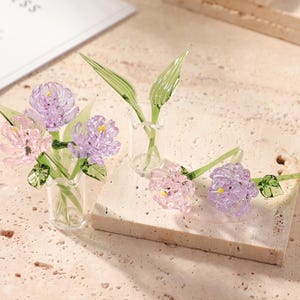 May include: Glass flower sculptures in a clear vase and on a stone block. The flowers are a mix of pink and purple with green stems and leaves. The glass flowers are delicate and detailed, showcasing a floral theme.