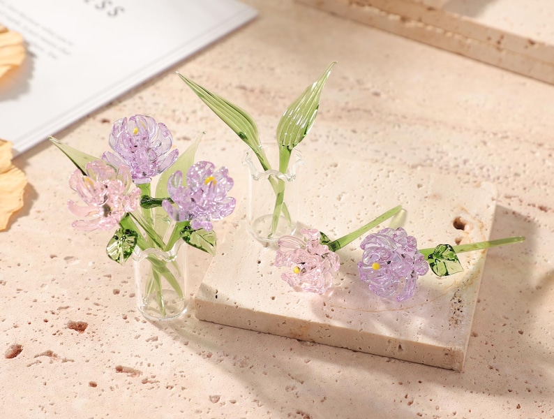 May include: Glass flower sculptures in a clear vase and on a stone block. The flowers are a mix of pink and purple with green stems and leaves. The glass flowers are delicate and detailed, showcasing a floral theme.