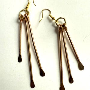 May include: A pair of handmade copper-colored earrings. Each earring features a gold-colored hook and a small ring connecting to three copper-colored rods. The rods have a teardrop shape at the end.