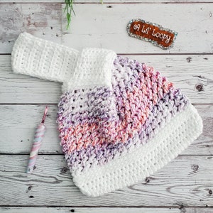 May include: A white crocheted hat with a pink and purple striped pattern. The hat has a wide brim and is made with a soft, textured yarn. The hat is sitting on a white wooden surface.