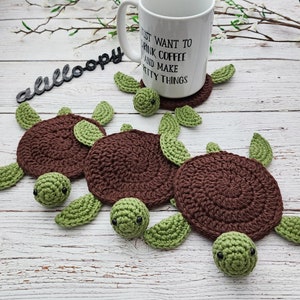 May include: Four crocheted turtle-shaped coasters in brown and green. The coasters are on a white wooden surface.